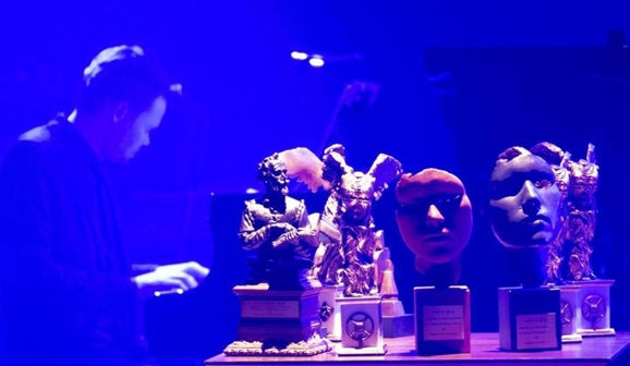 statueatki nagród w konkursie Debiutów stojące na stoliku. W tle jeden z laureatów gra na fortepianie